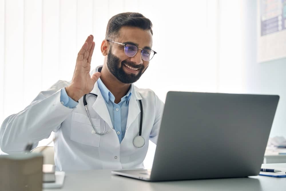 Male doctor smiling and waving toward laptop